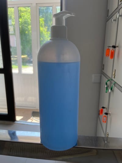 A close-up, eye-level, vertical medium shot captures a large, translucent plastic bottle filled with blue liquid, likely hand sanitizer, dominating the foreground. The bottle has a white pump dispenser on top. The bottle is positioned on a reflective metallic surface that runs horizontally across the bottom of the frame. Behind the bottle, to the left, is a dark-framed window with multiple panes, revealing a blurred background of green foliage bathed in sunlight, suggesting an outdoor setting. To the right of the bottle, a bank of gray lockers is visible, each with a silver handle and some with dangling keys or tags. Specifically, two orange tags and two green straps with keys are attached to the locker doors. The lockers appear to be in a communal area, such as a gym, school, or workplace, possibly in Nisporeni, Moldova, based on external information. The lighting is bright, indicating daylight, and the overall scene is clean and orderly. No individuals are present in the image.