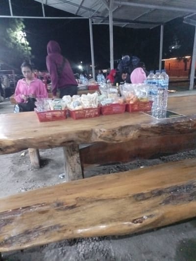 The image depicts a nighttime scene at a casual outdoor or semi-outdoor eating area, likely a food stall or warung in Pangalengan, Indonesia. The setting features sturdy, rustic tables and benches made from large, polished wooden slabs, resting on a gravel or dirt ground. An overhead shelter, constructed with metal poles and a corrugated roof, provides cover.
In the immediate foreground, a large wooden table is laden with multiple red plastic baskets, each filled with various pre-packaged snacks like chips and crackers. Several bottles of "AQUA" brand bottled water are also placed on the table.
On the left side of the table, a woman wearing a pink or magenta zip-up hoodie is seated, looking towards the camera with a neutral expression. A black bag is visible next to her. Standing behind her is a person in a dark purple or maroon hooded sweatshirt, facing away from the viewer. Further back, several other people are present; some are seated, including an individual in a red hoodie and another in a light pink top. Two figures in dark blue jackets or rain gear can be seen in the distant background.
The scene is lit by artificial lights, indicating the late hour. The activity appears to be casual dining or socializing at a food establishment. The overall atmosphere suggests a relaxed, informal gathering place. Aside from the "AQUA" branding, text on snack packages like "RINGAN" and possibly "BAGELEN" is faintly discernible.