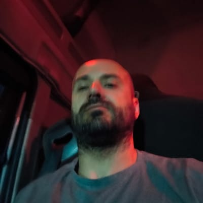 A close-up, blurry photo depicts a bald man with a beard wearing a gray t-shirt. The lighting is predominantly red, with some teal. The setting appears to be inside a vehicle, possibly a truck, with a dark interior and part of a driver's seat visible. The photo's angle is from below, looking up at the man. There is no clear indication of a location within Alcàsser, Spain, or any specific activity or interaction taking place.