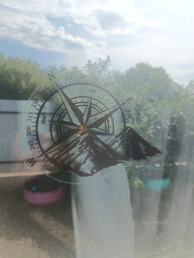 This media file features a close-up view of a circular sticker on a window. The sticker is a design of a compass with a mountain silhouette in the lower right portion. The compass has directional markings and degree increments, including "N", "ne", "nw", and "W", along with numerical values ranging from 15 to 345 degrees. The central part of the compass has a starburst design.
The background visible through the window suggests an outdoor scene. Green trees and foliage are prominent, with a bright, possibly sunny sky with scattered clouds overhead. In the lower left, a pink planter with a blue object is visible, and a glimpse of a fence can be seen. Further in the background, another planter, this one teal, is partially visible. The lighting and sky indicate it is daytime. The overall impression is of a sticker on a window looking out onto a natural or garden-like setting. The weather appears to be pleasant, with a bright sky. No people are visible in the frame.