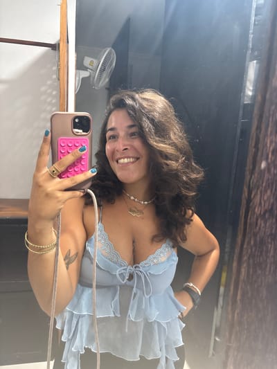 A young woman with curly brown hair takes a mirror selfie in what appears to be a changing room or bathroom, with a wall-mounted fan visible in the background. She is wearing a light blue, sheer ruffled top with lace trim, a pearl necklace with a gold pendant, and several bracelets. Her fingernails are painted bright blue, and she is wearing a gold ring on her right index finger. She is holding a rose gold phone with a pink PopSocket attached. A hummingbird tattoo is visible on her left forearm. The room has wooden accents, and dark curtains or doors are present to the right. The overall lighting suggests an indoor environment with some artificial light and possibly natural light. The specific location in Khan Daun Penh, Cambodia cannot be determined from the visual evidence alone.