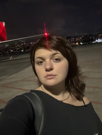 A person with shoulder-length brown hair and fair skin poses for a selfie at night. A city skyline with blurred lights is visible behind them. They wear a black top with a chain necklace. A nose ring is also visible. A red light reflects in the corner of the frame. The background appears to be an airport tarmac.