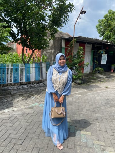 A forensic description of the scene in Ketegan, Indonesia, reveals a single adult female standing on a paved pathway under an overcast sky. The subject is of fair complexion, smiling and looking slightly to the right of the camera. She wears a light blue long-sleeved dress, an off-white crocheted sleeveless vest, and a light blue hijab covering her head. A gold-colored watch and bracelet are visible on her left wrist. She carries a light brown handbag with a decorative chain accessory over her left forearm, and wears light pink open-toed sandals.
The pathway consists of interlocking grey and blue pavers. To the subject's left, a low fence with blue and white vertical slats fronts a patch of green plants, likely cornstalks. Behind these is a reddish-orange building with a single window, partially obscured by a large leafy tree. To the subject's right, a simple building with a corrugated roof features a section painted with red and white vertical stripes. An improvised streetlight pole, supporting a dark lamp fixture, leans near this building. A sign partially obscured by foliage advertises "Bu Kurnia Catering & Snack Box" on the building's right side, surrounded by potted plants. Another large tree is present on the far right.