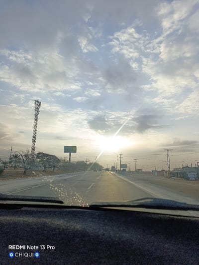 The image captures an asphalt road scene in La Cruz del Pastel, Venezuela, at either sunrise or sunset, viewed from within a vehicle. The sun, positioned low on the horizon and slightly right of center, emits intense light, causing a prominent starburst lens flare and numerous bright, specular reflections on the windshield, indicative of water droplets or dust.

The sky above is predominantly covered by light grey and white stratocumulus clouds. In the middle ground, the road, appearing to be a two-lane thoroughfare, stretches into the distance. To the left, a tall, lattice-structure cellular communication tower stands prominently, followed by a dry, sparsely vegetated landscape featuring trees and a large, unreadable billboard. To the right, a line of utility poles with power lines extends down the road, with indistinct low-rise structures and blurry human figures discernible on the roadside further ahead. Several vehicles are visible on the road in the distance.

The immediate foreground displays the dark, textured surface of a vehicle's dashboard and the base of the windshield. A digital watermark "REDMI Note 13 Pro" and "CHIQUI 🧿🧿" is present in the lower-left corner. No direct interactions between people are discernible.