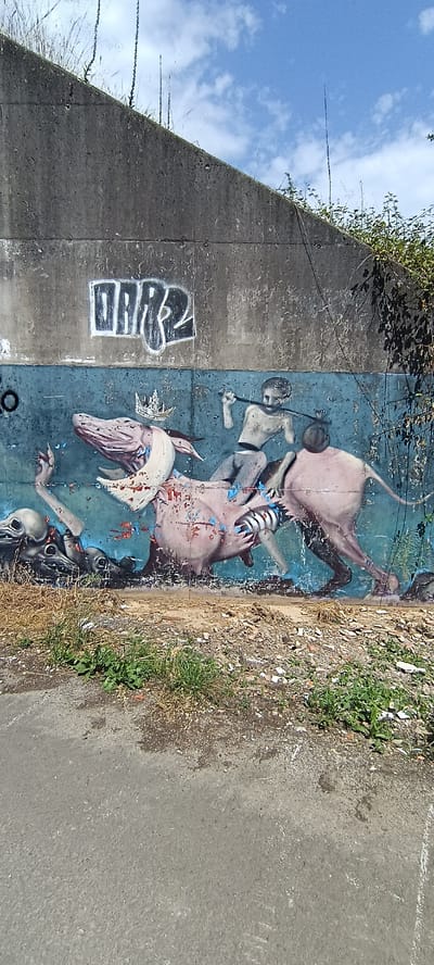 The image captures a section of a weathered concrete wall in Siero, Spain, adorned with graffiti. The wall, showing signs of age and damage, features a large mural painted on its lower half. The mural depicts a surreal scene with a pink, amorphous creature with a crown on its head, seemingly being ridden by a headless figure holding a stick. The creature has multiple disembodied heads emerging from its body, with sharp claws. To the right of the creature, another pink, fleshy form with a tail and visible legs is depicted. Splatters of red and blue paint are scattered across the mural. Above the mural, the graffiti tag "DMR2" is spray-painted in white and black. The upper part of the wall is covered by vegetation, and the sky above is blue with scattered clouds. The foreground shows a rough, paved surface with patches of dry grass and debris.
