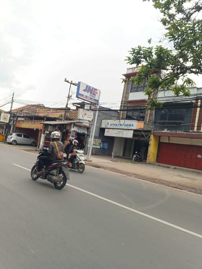 A street scene presents two people on motorbikes driving on the road. A JNE express office and Athaya Farma are visible, flanking a two-story building with closed red shutters and a yellow accent. A "Plosok Dele" restaurant can be glimpsed further down the street, where a white car is parked. Above, a cloudy sky sets the scene.