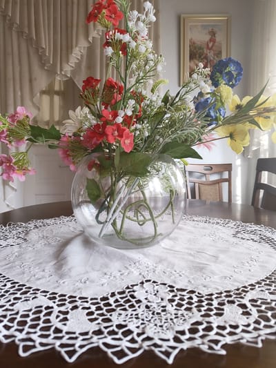 A still life arrangement features a large glass vase filled with a vibrant bouquet of artificial flowers. The flowers are a mix of colors including red, pink, white, yellow, and blue, with various types of blooms and greenery. The vase is placed on a circular, dark wooden table, which is partially covered by a white lace doily. The doily has intricate floral patterns and a scalloped edge. In the background, sheer curtains are drawn back, revealing a softly lit interior. A framed painting of a woman in a hat stands on the wall behind the curtains. Two wooden chairs with woven seats are visible in the middle ground, suggesting a dining or living area. The overall impression is one of a tastefully decorated indoor space, likely a home in Recea, Moldova. The lighting suggests it could be daytime, possibly morning or afternoon, with diffused light coming through the windows. No activity is taking place; it is a static composition. There is no visible text in the image.