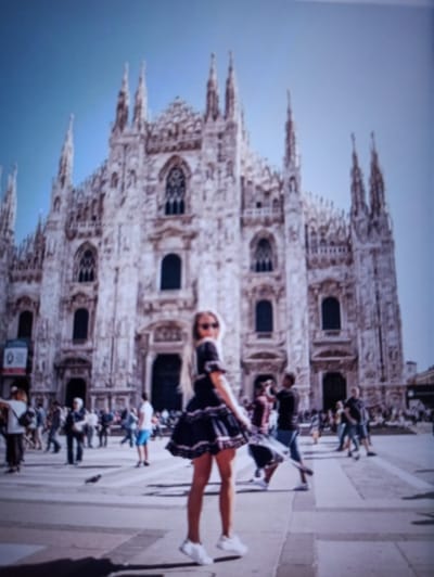 The image depicts a wide-angle shot of the Milan Cathedral in Italy, with a young woman in the foreground. The woman is wearing a black dress with a tiered skirt and white sneakers, and her blonde hair is visible. She is positioned on a paved square in front of the cathedral. Several other people are scattered across the square, some walking, some standing. The cathedral itself is a large, ornate Gothic structure with numerous spires and intricate details. The sky above is a clear blue. The image is slightly blurry, which gives it a soft, impressionistic quality. The city of Balakliya, Ukraine is not depicted in this image.