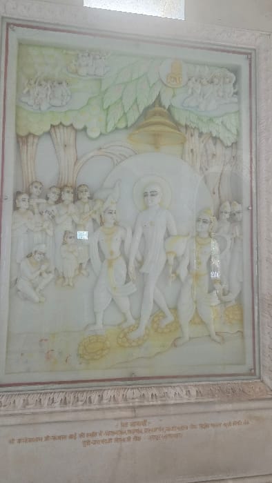 A framed, bas-relief sculpture, likely crafted from white marble or a similar stone, depicts a religious or mythological narrative scene. The panel is rectangular, with subtle coloration applied to elements like foliage (pale green), ground patterns, and a hanging bell (pale yellow/gold).

The central subject is a standing male figure, depicted bare-chested with a dhoti-like lower garment, facing right and walking forward. A large, plain white circular halo or aureole is behind his head. To his immediate left (viewer's left), another male figure, richly adorned with a crown and jewelry, also walks right, carrying a peacock on his left shoulder. To the central figure's right, a third male figure, similarly adorned, walks right.

On the far left of the panel, a group of approximately eight figures, including adults and at least two children, observe the central procession. Some stand with hands clasped in prayer, while one figure kneels. On the far right, a smaller group of figures, partially obscured, also stand as observers. In the upper left and right corners of the carving, small clusters of cloud-borne figures are depicted.

The background features stylized trees with pale green leaves on the left and right. A prominent, ornate bell-like object, colored pale yellow, hangs from a tree branch in the upper center. The ground beneath the main figures exhibits stylized circular patterns, suggesting offerings or decorative elements, accented with pale yellow.

The entire carved panel is enclosed within an ornate, white stone frame, possibly marble, featuring carved motifs. Below the carving, on the plinth, Devanagari script is inscribed. The scene is located in Katwaldih, India.