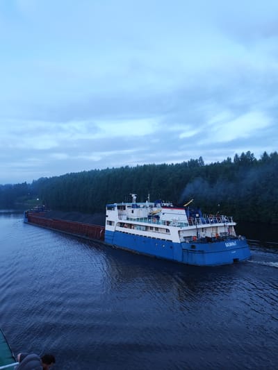 An overcast day in Volokov Most, Russia, shows a cargo barge laden with dark material moving down a wide, dark blue river. The barge is composed of a long, flat cargo area with red railings, followed by a white and blue superstructure with windows. Smoke billows from a chimney on the superstructure. The river is bordered by a dense, dark green forest. In the foreground, a person is leaning over the edge of what appears to be another vessel. The sky is a pale, hazy blue with wispy clouds.