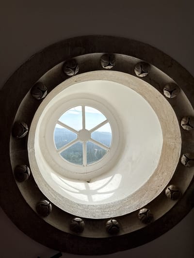 A circular window in the Quinta da Regaleira, Sintra, Portugal, is viewed from below. The window is framed by two concentric stone rings. The outer ring is decorated with evenly spaced sculpted bulbous shapes. The inner ring is smooth and white. The window itself has a simple white frame and six panes separated by white bars. Through the window, a landscape of green trees and hills can be seen under a pale blue sky with some clouds. Sunlight streams into the window, casting shadows on the interior.