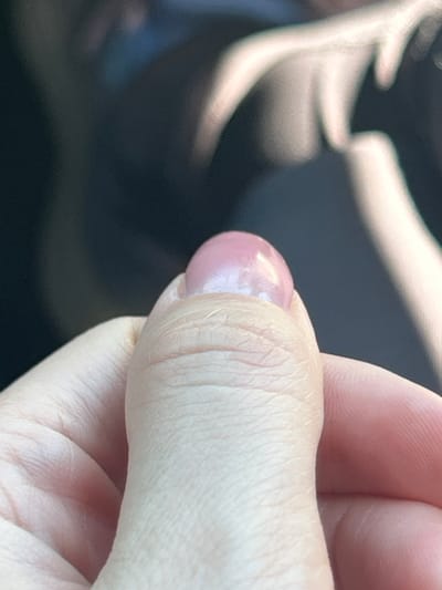 A close-up, eye-level, outdoor, daytime shot focuses on a person's thumb with a pink, glossy nail. The thumb is positioned in the lower center of the frame, with skin texture clearly visible. The background is blurred and dark, with streaks of light suggesting movement or a reflective surface, possibly the interior of a vehicle. The scene is set in Everett, United States of America.