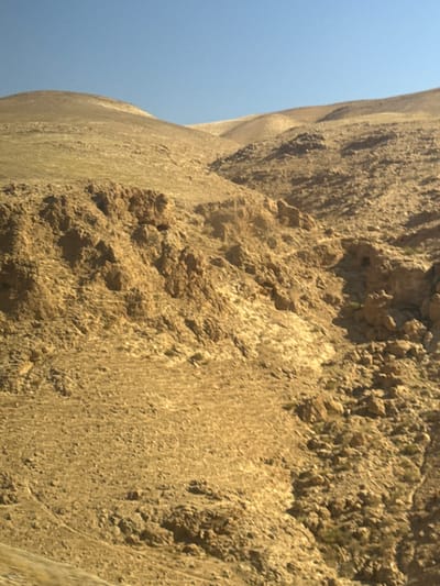 The media file is a photograph of a steep, rocky cliff face with a clear blue sky in the background. The cliff is composed of rough, textured rock in shades of brown and tan, with areas of shadow and highlights suggesting uneven terrain. Some patches of green vegetation are visible on the cliff face, indicating a dry environment.
The perspective is from below, looking up at the cliff. The rock appears to be weathered and eroded, with many small stones and debris scattered across its surface. The curve of the cliff suggests a large, natural formation, possibly a canyon or a mountainside.
The bright blue sky indicates a clear day with ample sunlight, likely midday or afternoon given the strong contrast and shadows. There are no people or man-made objects visible in the frame, focusing solely on the natural landscape. The overall impression is one of rugged, natural beauty and perhaps a sense of scale and grandeur due to the dominance of the cliff in the composition.
The location context of Arad, Israel, suggests a desert or arid environment, which is consistent with the appearance of the cliff and vegetation. The media file captures a static scene of a geological formation under bright daylight conditions.