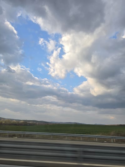 The image displays a rural landscape, observed from a moving vehicle on a highway in Yazdach, Bulgaria. The foreground features a three-tier galvanized steel highway guardrail positioned horizontally across the lower third of the frame, with a section of dark grey asphalt road visible beneath it. Beyond the guardrail, an expansive green field stretches across the midground. A line of trees or shrubs with light-colored, possibly flowering foliage, borders the field further back. In the background, a series of undulating hills or low mountains are visible. The sky occupies the upper two-thirds of the image, dominated by a mix of light grey and white stratocumulus clouds. Irregular patches of clear, light blue sky are discernible, particularly in the upper-left and central-right areas. No people, active subjects, or discernible interactions are present. The scene is illuminated by diffused natural daylight.
