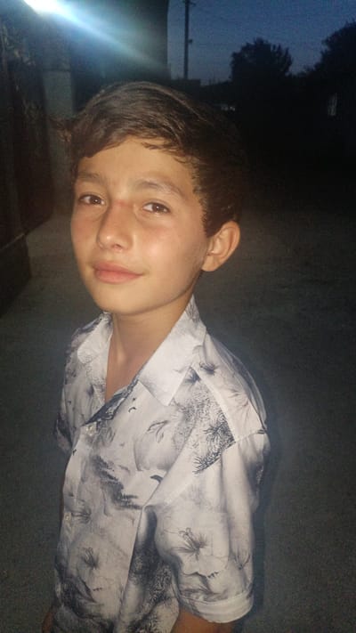 A medium shot shows a young boy with dark hair and brown eyes, looking towards the camera. He is wearing a short-sleeved, collared shirt with a black and white floral pattern. The shirt is buttoned at the top. His skin appears tanned, and there is a small red mark on his forehead just above his right eyebrow. The boy is positioned in what appears to be an outdoor setting, possibly at dusk or in low light conditions, as indicated by the dim background and the presence of a bright light source visible in the upper left corner of the image. Trees and a utility pole are silhouetted against the dark sky. The ground is visible in the foreground and appears to be paved. The scene is consistent with a residential or rural area in Tajikistan, given the location context. No text is visible in the image.