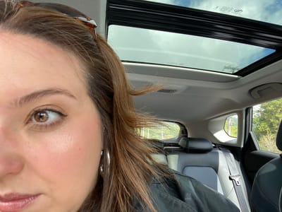 A close-up, canted-angle photograph shows a woman's face occupying the upper right portion of the frame. She is inside a vehicle, evident from the car interior, including the sunroof, seats, seatbelt, and headrest, visible in the background. Outside the car windows, trees are visible. The location is presumed to be Nova Petrópolis, Brazil, but this is not confirmed within the image itself.