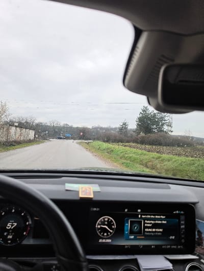 The image provides a driver's perspective from within a vehicle on a cloudy day in Gorski Izvor, Bulgaria. The foreground shows the vehicle's dashboard, with a partial view of the steering wheel on the left. A digital speedometer on the dashboard displays "57". The central digital display indicates the time as "09:21", the date as "22.03.2024", and an ambient temperature of "26°C". This display also shows "Here's BGM for work / Music Player" and the track "YOUNG BY YOUNG Featuring a Krosno", with "-3:12" remaining. A small, rectangular, framed object rests on the dashboard surface.

The view through the windshield shows a paved road extending forward, flanked by an aged, light-colored structure with visible damage on the left, and a grassy verge leading to a field with green vegetation and bare soil on the right. Sparse, leafless trees are visible behind the structure on the left. In the mid-distance on the right, a prominent coniferous tree stands, partially obscuring a large billboard structure. Further down the road, a blue vehicle is visible, positioned near what appears to be an intersection or bend, with a circular red sign to its right. The sky is uniformly overcast. No individuals are depicted.