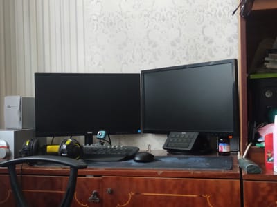A desk with two computer monitors is set up against a wall with striped and floral wallpaper. On the left monitor, there is a webcam. A black keyboard is in front of the left monitor, and a black mouse is in front of the right monitor. To the left of the monitors, there are two gray boxes and a pair of black and yellow headphones. On the right side of the desk, there is a black keypad and a small bottle with a blue and white label. A black office chair is in the foreground. The scene appears to be indoors, likely in a home or office.