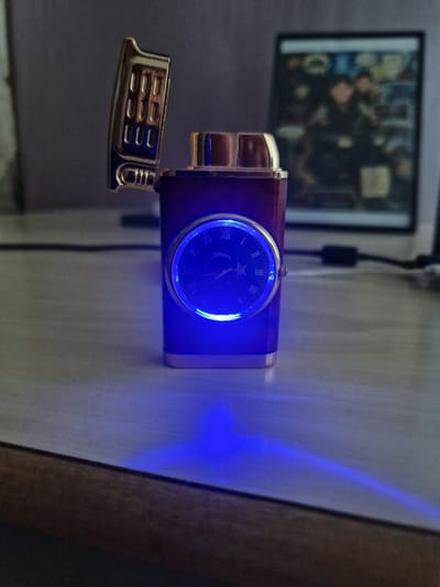 The image shows a close-up of a gold and brown cigarette lighter with a clock face. The lighter is open, revealing a metallic, ribbed bottom section. The clock face is circular, set into the side of the lighter, and illuminated by a bright blue light, casting a blue glow onto the surrounding wooden surface. Roman numerals mark the hours on the clock face, and a star symbol is visible at the 12 o'clock position. The hands of the clock appear to be pointing to approximately 10:15. In the background, slightly out of focus, is a framed picture on a wall, depicting what appears to be a group of people in a dimly lit setting. The overall scene suggests an indoor, possibly domestic, setting, with the focus on the unique lighter. The blue light from the lighter is a prominent feature, creating a modern and somewhat dramatic effect against the darker tones of the lighter and the wooden surface. No specific time of day or weather conditions can be determined. The location context is Rikava, Latvia.