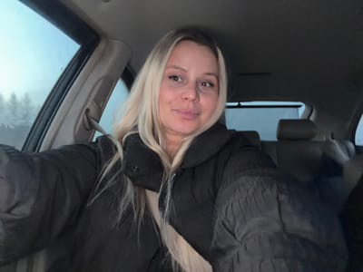 A blonde female is seated within the interior of a passenger vehicle, appearing to take a selfie. She wears a dark, possibly black, puffer jacket over a light-colored garment, with a light-colored seatbelt diagonally across her chest. Her expression is a slight smile, and she looks directly at the camera. The vehicle's interior features dark seating and a dark headliner. Through the left-side window, an overcast or dim sky in shades of light blue-grey is visible, with silhouettes of trees in the background. The scene is located in the city of Gol'yany, Russia.