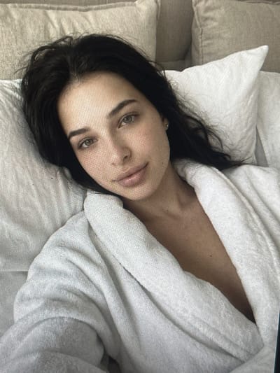 The image shows a close-up, head-and-shoulders view of a young woman with dark hair, wearing a white bathrobe, lying in bed. She is looking directly at the camera with a neutral expression. The background consists of pillows and what appears to be a headboard. The image quality is grainy, suggesting it may be a low-resolution photo or a screenshot. The location of Pruszcz Gdański, Poland is not ascertainable from the visual information provided in the image.