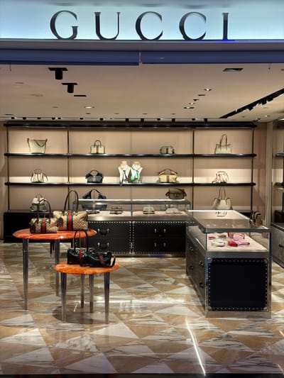 The image shows the interior of a Gucci store. A vertical "GUCCI" sign in large, sans-serif letters is positioned on the left. A display of Gucci handbags is on the adjacent wall. These bags are arranged on shelves. In front of the shelves, there are display tables holding more bags. The floor has a marble-like design. The location is Taoyuan City, Taiwan.