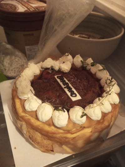 The scene depicts a round baked cheesecake or tart, approximately 20-25 cm in diameter, resting on white paper within what appears to be a refrigerated environment in B. Nam Ngao, Laos. The cake features a golden-brown crust and is topped with a dark reddish-brown fruit compote or jam. Around the perimeter of the topping, 15 individual dollops of white whipped cream or frosting are arranged, each adorned with a small sprig of green herbs, possibly thyme or mint. A rectangular white icing plaque, inscribed with the dark lettering "DRCYMBO," is centrally embedded in the fruit topping. In the background, to the left, a large brown cylindrical container, possibly for ice cream, with visible partial text including "CHOCO," is present. A translucent plastic bag hangs behind the cake towards the center. To the right background, a light-colored ceramic bowl with subtle horizontal banding is visible. The foreground and the surface directly beneath the cake appear to be a metallic or reflective shelf. No individuals are present.