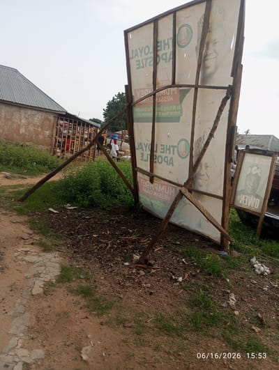 In the foreground of the image is a dirt road with a cracked concrete path on the left. To the right, the ground is covered in dark soil and debris, with patches of green grass. A large, partially collapsed billboard is the central focus. The billboard features political posters, including one with a man in a patterned hat and the text "VOICE OF HOPE OWEN". Another poster on the billboard shows a smiling man in a patterned hat, with the text "THE LOYAL APOSTLE". The billboard is supported by wooden poles, some of which are broken or tilted, indicating it is unstable. In the background, there is a building with a corrugated metal roof, and further back, more greenery and trees. Several people are visible in the middle ground, some near a wooden structure that appears to be a market stall. One person is carrying a blue bucket on their head. The overall scene suggests an outdoor environment in a semi-urban or rural area, possibly a market or roadside. The date and time stamp "06/16/2026 15:53" is visible in the bottom right corner.