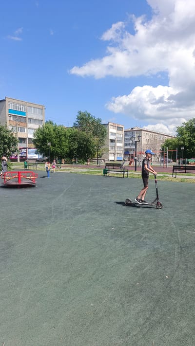 The image depicts a sunny day in a city park in Kuybyshev, Russia, likely during summer. A young boy wearing a blue baseball cap, black t-shirt, and black shorts is riding a black electric scooter on a green rubberized surface. He is positioned in the middle right of the frame. In the background, several residential buildings and trees are visible. To the left of the frame, a red and yellow merry-go-round and a blue spring rider are present on the playground. Other children and adults are scattered in the background, some near playground equipment or on benches. The sky is a clear blue with scattered white clouds.