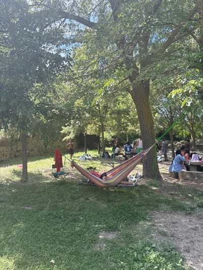 The image depicts a leisurely outdoor gathering in a park-like setting. A colorful hammock is strung between two trees in the foreground, with a person lying in it. Several other individuals are dispersed throughout the scene, some sitting at picnic tables, others standing and conversing. The environment is characterized by lush green grass, mature trees providing ample shade, and a stone wall in the background. The presence of picnic tables and people in casual attire suggests a recreational activity such as a picnic or a day out. The location is identified as Campezo / Kanpezu, Spain.