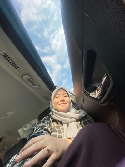 A woman wearing a light gray hijab and a plaid shirt is sitting in a car, looking up towards the sunroof. The sunroof is open, revealing a bright blue sky with scattered white clouds. The car's interior is visible, including part of the door handle and air vents. The woman has a light gold ring on her left ring finger. The scene appears to be outdoors, possibly during the daytime, under a sunny sky. The location of Sungai Lakum, Indonesia is not specified in the image or its surrounding context.