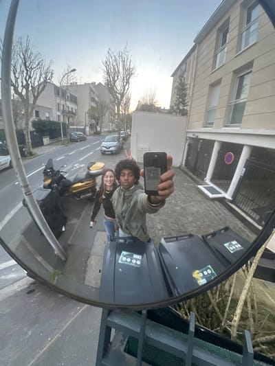 A wide-angle, distorted image captured via a convex traffic mirror depicts two individuals on a street in Le Perreux-sur-Marne, France. In the foreground, a male subject with dark, curly hair, wearing a light olive-green jacket, extends his right arm holding a black smartphone directly towards the mirror, acting as the photographer. To his left, slightly behind him, a female subject with long, dark hair smiles and displays a 'peace' sign (V-sign) with her right hand; she is attired in a dark, long-sleeved top and light-colored jeans. Reflected in the lower portion of the mirror are three dark grey waste bins, each bearing green and white informational stickers. The background reveals a paved street scene: a two-lane road with a double white line, a yellow and black scooter parked on the sidewalk, and several parked cars, including a silver Renault Clio. Multi-story residential buildings with various windows and balconies line the street. Bare deciduous trees are visible, and a red circular 'No Parking' sign with a diagonal line is affixed to a building wall. The sky appears clear and bright, indicating daytime.