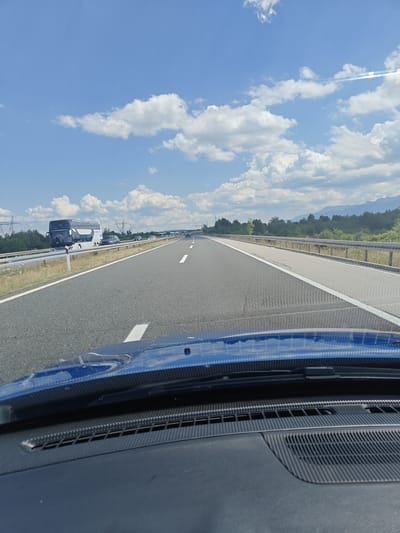 The image shows a view from inside a vehicle driving on a highway on a sunny day. The road is asphalt with white lane markings. In the distance, mountains and trees are visible under a blue sky with scattered white clouds. A double-decker bus and several cars are also present on the highway. The foreground shows the dashboard and windshield of the vehicle. The location is identified as Barlete, Croatia.