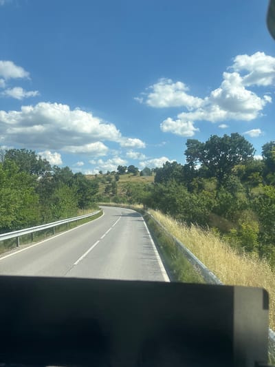 The image shows a view from a vehicle traveling on a paved road. The road curves to the right, with white lane markings visible. On the left side of the road, there is a metal guardrail and lush green trees. The right side of the road features a similar guardrail and tall, dry grass. Rolling hills covered in trees and vegetation extend into the background. The sky is a vibrant blue with scattered white cumulus clouds. The bottom of the image is obscured by a dark, blurry object, likely part of the vehicle's interior. The scene is consistent with a rural or suburban area, possibly in or around Mladinovo, Bulgaria, given the natural landscape and vegetation. No people are visible in the image.