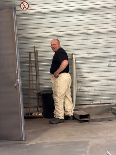 A man is lying on the ground in what appears to be an industrial setting. He is wearing a black t-shirt, light-colored pants, and dark boots. His head is visible, and he is looking towards the camera with a neutral expression. In the background, there is a corrugated metal wall, a concrete floor, and some metal shelving units. There is also a circular sign with a red border and a diagonal line through a cigarette, indicating "no smoking." The overall scene suggests an indoor, possibly storage or workshop area, with utilitarian elements. The lighting is consistent with indoor illumination, and there are no clear indicators of time of day or weather conditions.