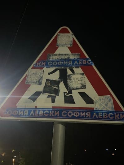 The image shows a nighttime view of a pedestrian crossing sign in Gorna Malina, Bulgaria. The sign is triangular with a red border and a white interior, depicting a black silhouette of a person walking across white stripes. The sign is covered in various stickers and graffiti. Two horizontal blue bands run across the sign, the top one bearing Cyrillic text that appears to read "СОФИЯ ЛЕВСКИ" (Sofia Levski) and the bottom one also displaying "СОФИЯ ЛЕВСКИ" in red letters on a blue background. There are also smaller, square-shaped stickers or graffiti elements scattered across the sign, some appearing as abstract patterns or scribbled text. The sign is mounted on a metal pole. The background is dark, suggesting it is nighttime, with faint lights and a power line visible in the upper left corner.