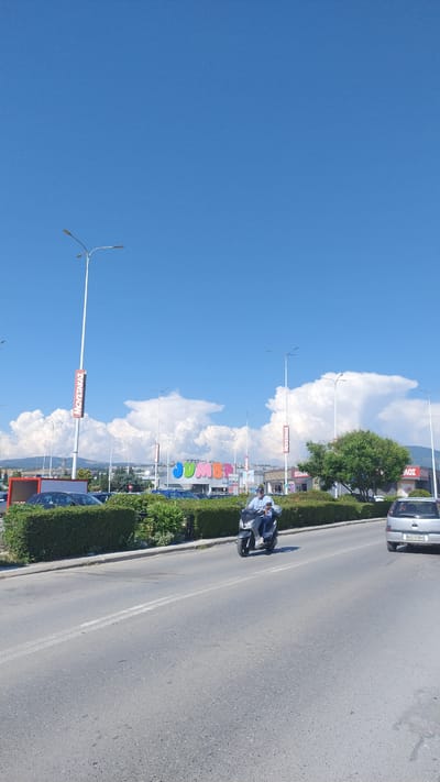 This image, taken in Pylaia, Greece, shows a street scene on a clear, sunny day. The sky is a vibrant blue, dotted with fluffy white clouds. On the right side of the frame, a paved road runs alongside a neatly trimmed green hedge. Cars are parked along the roadside, and a silver car with license plate "NKE-5964" is visible. A man on a scooter, wearing a light blue shirt, rides on the road.

In the background, several tall lampposts stand against the sky. Further back, large, colorful signs are visible, one of which spells out "jumbo" in large, individual letters in green, blue, and red. Other signs with red lettering read "MOVETAKAE," which appears to be Greek text. A few trees, including a large one with green foliage, add to the scenery. The overall impression is a bright and active commercial area on a pleasant day. The orientation of the image is sideways.