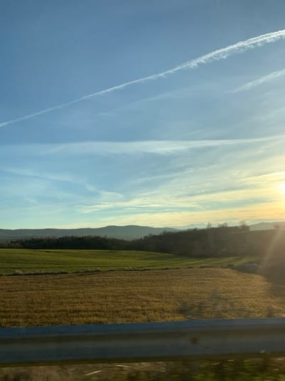 The image captures a serene landscape from a roadside perspective in Grevena, Greece, under a vast sky, likely at sunrise or sunset. The upper portion of the frame is dominated by a clear blue sky, gradually shifting to a bright golden glow in the upper right corner where the low sun creates a gentle lens flare. Streaks of wispy white clouds extend horizontally across the sky, interspersed with distinct, linear contrails from an aircraft, with one prominent contrail running diagonally from the upper left.

Below the expansive sky, a range of distant, undulating hills or mountains forms a dark, hazy blue-green silhouette across the mid-ground, stretching towards the horizon. In the foreground, below the mountains, are agricultural fields. The field closest to the hills appears a lush light green, possibly indicating young crops, while the field occupying the immediate right foreground is a contrasting golden-brown, suggesting drier vegetation or harvested land. A sturdy, silver-grey metal guardrail runs vertically along the far right edge of the image, implying the view is taken from beside a road or from a vehicle. No people or visible text are present. The scene conveys a sense of peaceful rural scenery under a dramatic sky.