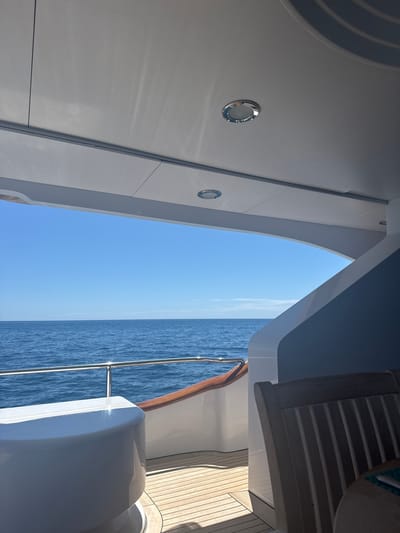 The image shows a view from inside a yacht, looking out through an opening. The left side of the frame is dominated by a white wall with two round, chrome light fixtures. The opening reveals a view of the ocean and a clear blue sky. On the right, there is wooden decking and part of the yacht's exterior, including a railing and what appears to be a white structure. The location is an unidentified point off the coast in Mexico, based on the landscape.