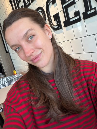 A woman with long brown hair and green eyes is pictured wearing a red and brown striped shirt. She is smiling slightly at the camera. She has fair skin and a small stud earring in her left ear. The background includes a wall with black and white tile. There is also writing on the wall in black lettering. The location may be in the city of Ub Municipality, Serbia.