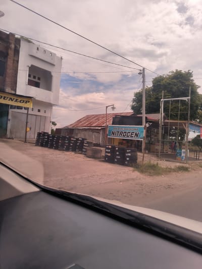 The image presents a daytime street scene in Meukokuthang, Indonesia, captured from the perspective of someone inside a vehicle, with the car's dashboard visible at the bottom. The setting appears to be a commercial roadside area.

On the left, a two-story building features a white, modern upper section with small windows and a darker, possibly brick, ground floor. A yellow "DUNLOP" banner hangs from the lower part of this building. In front, along the road, numerous stacks of black tires are neatly arranged, suggesting a tire sales or service business. A concrete basin or trough is positioned near some of these tire stacks.

Moving towards the right, a building with a reddish-brown corrugated metal roof is visible. Next to it, a larger, open-sided structure serves as another commercial outlet, likely a tire shop or service station. A prominent blue banner on this structure reads "BAN BARU BAN SECOND" (New Tires, Used Tires) and "NITROGEN" in large white letters. Below, smaller text advertises services including "ISI PULSA" (top-up credit), "BAYAR LISTRIK" (pay electricity), "PDAM" (water utility), "BPJS" (social security), "OJEK" (motorcycle taxi), and "GRABPAY," with the word "Pojok" (corner) also present. Additional stacks of tires are visible around this area. A metal frame structure stands to the right of this establishment.

A wooden utility pole with power lines extends upwards on the right side of the frame, supporting a street light. Behind the roadside structures, a large, dense green tree is prominent. The sky is mostly overcast with scattered white and grey clouds, suggesting either late morning or early afternoon. There are no discernible people engaged in specific activities, and the scene depicts a static snapshot of the roadside environment.