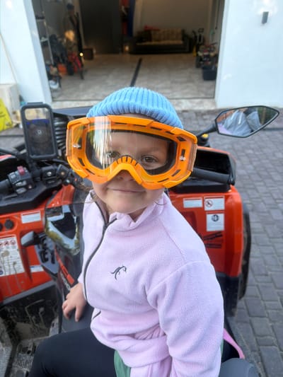 A young person wearing orange ski goggles and a light blue beanie is sitting on an orange ATV. The person is also wearing a light pink fleece jacket with a small black deer logo on the left side of the chest and black leggings. The ATV appears to be in a somewhat muddy or dirty outdoor setting, possibly a driveway or yard, with brick paving visible. In the background, there's a glimpse of a garage or building with various items stored inside. The image is taken from a slightly low angle, looking up at the subject. The lighting suggests it's daytime. The person's eyes are visible through the goggles, and they appear to be looking towards the camera with a neutral expression. Text is visible on the ATV, including "FORCE" and possibly a model number like "1600". The reflection in the mirror of the ATV shows a gray sky and possibly trees. The image appears to be taken in Колюткино, Russia.