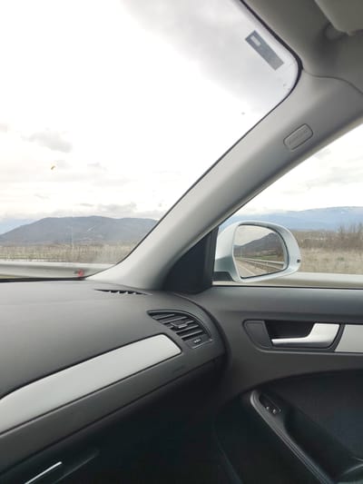 The image shows an interior view from inside a car, likely on a highway. The dashboard is visible, along with the passenger-side window and side mirror. The side mirror reflects the road and landscape that is visible out of the windshield. The landscape includes mountains and roadside vegetation. The sky is overcast. The location is Novachene, Bulgaria.