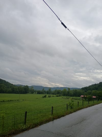The image depicts a rural landscape under an overcast sky. In the foreground, a paved road curves to the right. A wire fence runs along the edge of the road, separating it from a lush green field. Beyond the field, a line of trees forms a border against rolling hills. In the distance, nestled among the trees, are two small buildings with red roofs. The scene appears to be in a countryside setting, possibly in or near Stanton, United States, given the rural topography and architecture. There are no people or animals visible in the image. The sky is filled with thick, gray clouds, suggesting a possibility of rain or a recent downpour, as the road appears damp. The overall impression is one of quiet, natural beauty.