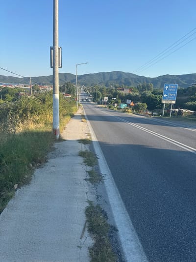 A road in Toroni Municipal Unit, Greece, stretches into the distance under a clear blue sky. On the right side of the road, a blue directional sign points to "Sykia" (up) and "Sarti" (left), with a "100m" indicator below. The road itself has two white lines marking its center and a paved sidewalk running alongside the left edge, bordered by grass and vegetation. In the background, green hills and scattered buildings are visible. A tall pole with a rectangular fixture stands on the left side of the sidewalk. Several cars are visible driving on the road in the distance. The overall scene depicts a typical rural or semi-rural road setting.
