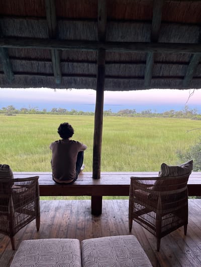 The image shows a person sitting on the edge of a wooden porch, looking out onto a grassy plain under a blue sky, seemingly in Khwai, Botswana. The porch is furnished with a wicker chair and a patterned sofa. The porch is covered with a thatched roof, supported by wooden beams. The person is dressed in a light-colored shirt and dark pants, with their back to the camera.