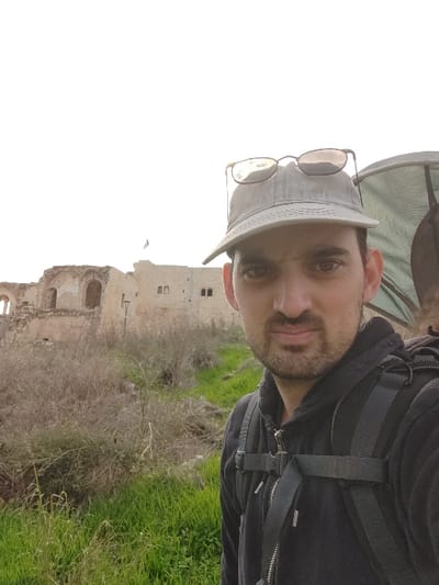 A male individual is positioned in the foreground, slightly to the right, in an outdoor setting identified as the Antipatris Fortress (Tel Afek) in Tel Afek National Park, Rosh HaAyin, Israel. He is seen from the chest up, wearing a dark hooded sweatshirt and a light grey baseball cap with dark sunglasses resting on the brim. A dark backpack with visible straps is on his back. He has short, dark hair, dark eyes, and stubble on his face, looking directly at the camera with a neutral expression. Behind him, to the left and extending across the midground, are the stone ruins of the fortress, characterized by arches and multi-story sections. A dark blue flag is visible atop a flagpole on the fortress's highest point. The terrain in the foreground and midground consists of a grassy slope with both vibrant green and dry, brown vegetation. The sky is bright white, suggesting an overcast day.