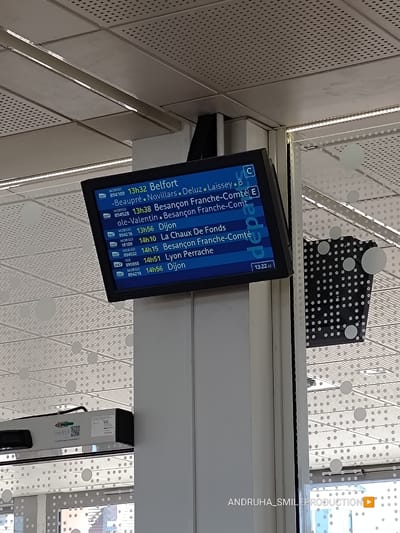 The image shows a departure board at a train station. The board displays train information, including departure times, destinations, and train numbers. The language on the board is French. The text "departs" is visible on the right side of the board. The time displayed on the board is 13:22:02. In the bottom right corner of the image, there is a watermark that reads "ANDRUHA_SMILEPRODUCTION" with a play button icon. The train station appears to be modern, with a tiled ceiling and glass partitions. There are no people clearly visible in the image.