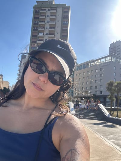 A woman in sunglasses and a Nike cap poses for a selfie. She is wearing a blue top. Tall buildings stand in the background under a bright sky. A group of people are also visible behind a railing. The image is slightly angled, giving a dynamic perspective.
