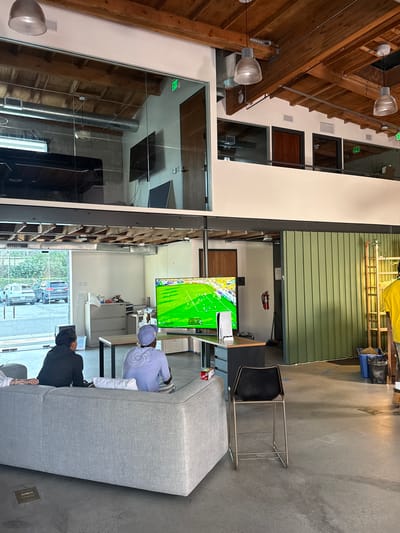 The image depicts an indoor space in what appears to be an office or collaborative workspace in Culver City, United States of America.
A TV screen displays a soccer match.
Two people are sitting on a gray couch, and one is watching the television.
Architectural details include exposed wooden beams, large windows offering a view of a city street, and industrial-style lighting fixtures. The overall environment suggests a modern, open-concept workspace.